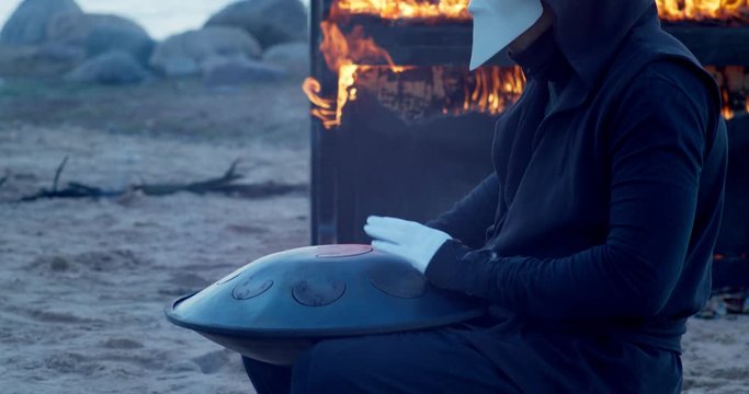 a man in a venetian mask on the seashore plays the glucophone against the background of a burning old piano