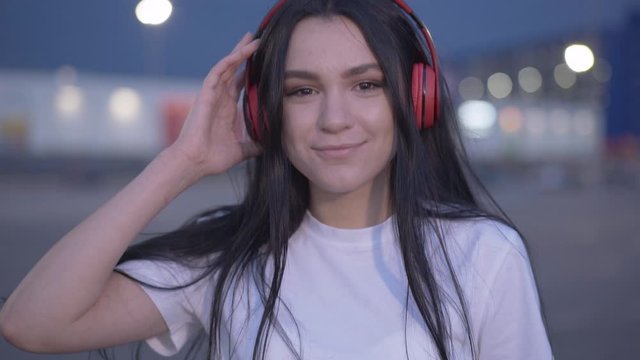 Camera goes from female feet in rollers to smiling face of beautiful young woman looking at camera. Portrait of positive skater standing at dusk in city. Happy woman skating in the evening.