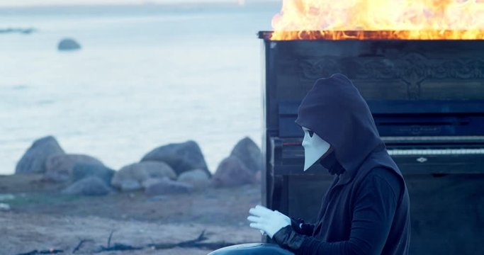 a man in a venetian mask on the seashore plays the glucophone against the background of a burning old piano