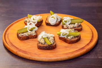 slices of black bread with meat pate decorated with pickles and onions on a round wooden tray. Close view