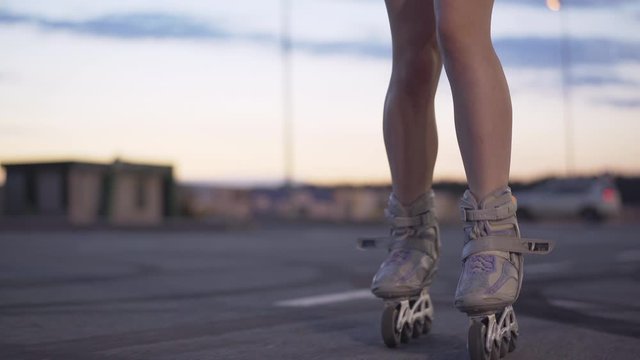 Slim female legs in roller skates riding at dusk on empty parking lot. Unrecognizable young Caucasian woman skating in the evening in city. Concept of hobbies and leisure.