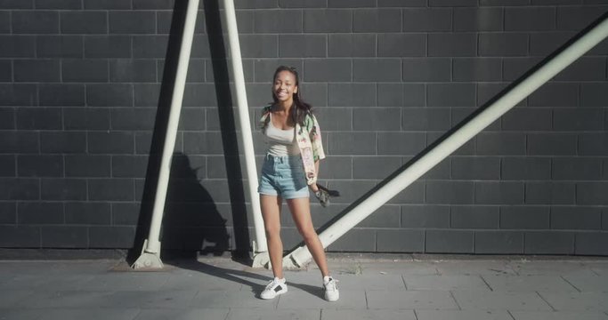 Happy funny mixed race female standing by urban wall holding penny skateboard outside, cheerful teen girl in trendy summer outfit looking to camera smiling positive moment carefree feeling of freedom