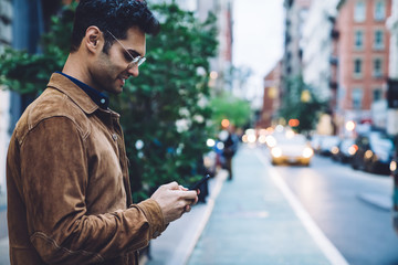 Cheerful ethnic young man in jacket texting on street