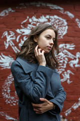 Fototapeta premium young thoughtful pretty girl in gray coat and turtleneck poses in front of red brick wall with graffiti. Street style portrait of female, urban photoshoot of elegant model