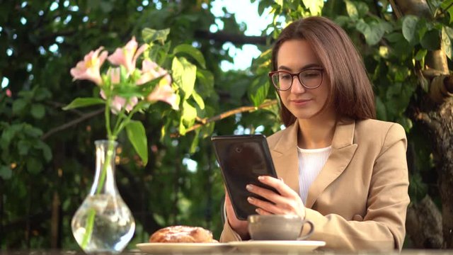 Young business woman sits in a cafe and writes text messages on a tablet. Girl with coffee and a bun on the summer veranda.