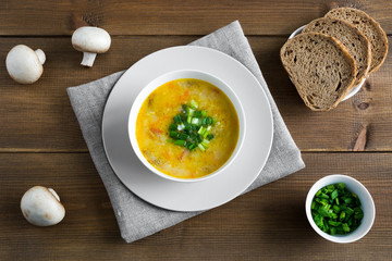 Champignon soup in white bowl on napkin. Whole champignons, bread flat lay with place for text on wooden brown background. Top of soup is decorated with green onion.
