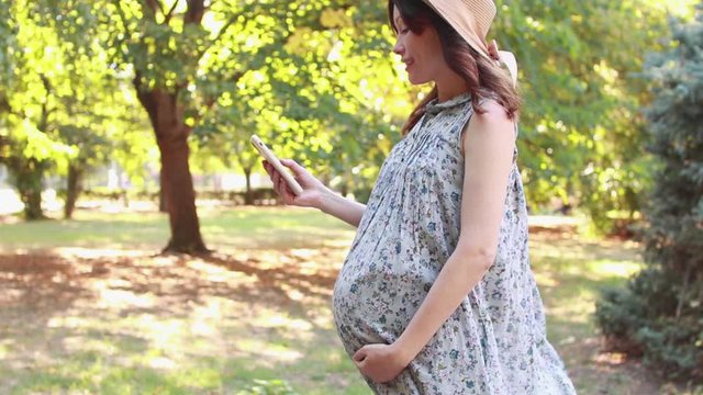 Pregnant woman using phone in the park on a summer day and stroking her tummy. Pregnancy and maternity concept. Place for text. Selective focus.