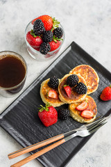 Cottage cheese pancakes with blackberries and strawberries on a dark plate with cutlery and a glass with coffee on a light background