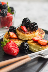 Cottage cheese pancakes with blackberries and strawberries on a dark plate with cutlery on a light background close-up