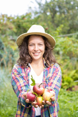 Smiling young woman holding in front of her heap of freshly picked red apples harvested by herself in an orchard garden. Happiness for ripe fruits of her family's labor. Farming concept. Vertical.