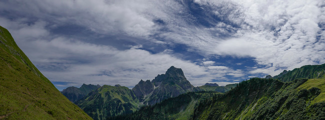 Panorama grüne Berghänge in den Alpen mit faszinierenden Wolken am blauen Himmel