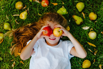 Young girl laying on green grass among pears, apples and small orange leaves.
