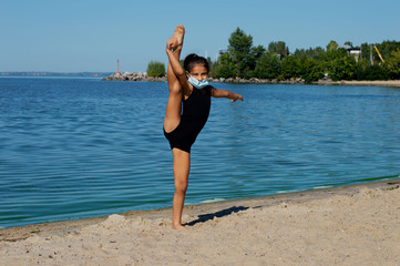 A gymnast girl in a protective medical mask, dressed in a sports swimsuit, performs gymnastic...