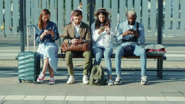 Serious multiethnic young travellers men and women sitting on bench at bus stop use phone together street stylish summer sunny tourists outdoors slow motion