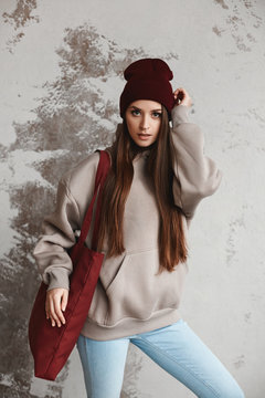 A young woman in a sweatshirt and modish hat posing with shopper next to the wall