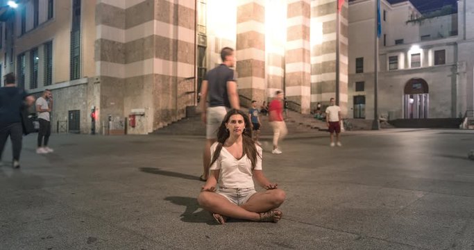 A 4K video time lapse of girl sitting cross-legged in the town square with background of boys playing soccer. Street Boys. Urban lifestyle concept.