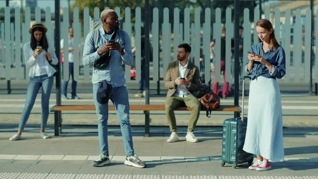 On bus stop mixed-races young males and females people standing keeping safe social distance use phone travelling street african american waiting outside vacation tourists slow motion