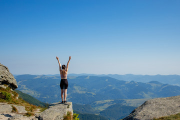 The young girl at the top of the mountain raised her hands up on blue sky background. The woman climbed to the top and enjoyed her success. Back view