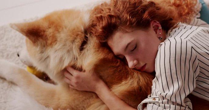 Tired young woman with red curly hair lying with closed eyes on floor carpet, taking rest napping cuddling up with Akita Inu Dog. Human animal bond. Close up top view.