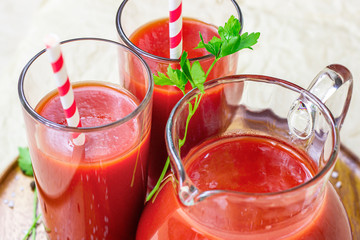 Fresh red tomato juice in a glass with a straw and jar with tomatoes on light wooden background.