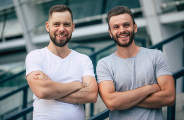 Two happy guys with crossed arms. Two young bearded handsome friends in t-shirts are posing together on the city background.