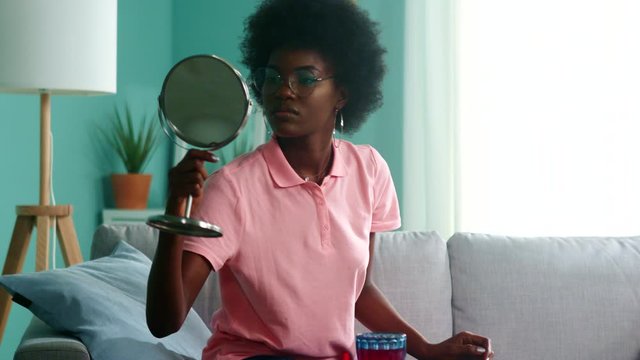 Young black woman, student, is sitting on sofa at home, getting ready to go outside, putting stylish glasses and looking at table mirror, beauty concept, Slow motion.