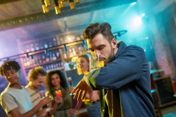 Handsome young man getting drunk, drinking a cocktail in the night club and friends chatting, having drinks at the bar counter in the background