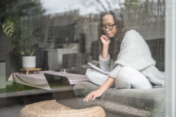 Asian woman wearing glasses smiles while working from home on her laptop computer, sitting on a sofa next to a patio door in the living room of a house in Edinburgh, Scotland, United Kingdom