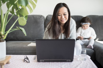Asian woman working from home smiles while typing on her laptop computer, sitting on a sofa in the living room of a house in Edinburgh, Scotland, United Kingdom, with her daughter playing beside her