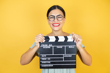 Young beautiful woman wearing glasses standing over isolated yellow background holding clapperboard...