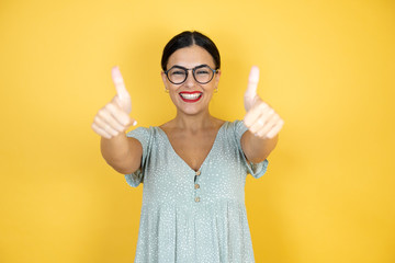 Young beautiful woman wearing glasses standing over isolated yellow doing happy thumbs up gesture with hands. Approving expression looking at the camera showing happiness background