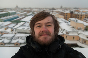 Portrait of a funny fair-haired boy with disheveled hair and a beard on the background of the snow-capped rooftops of St. Petersburg