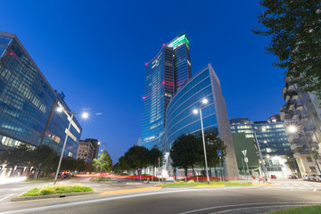 Milan, Italy - August 25, 2020: long exposition shot featuring a street view of Milan at dusk. Light trails are visible in twilight in front of Regione Lombardia headquarters skyscraper. - obrazy, fototapety, plakaty