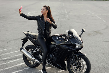 Obraz na płótnie Canvas Beautiful female biker makes selphie photo by smartphone while sitting on stylish sport motorcycle at outdoors parking.