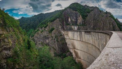 Panoramic photo of Vidraru dam or Barajul Vidraru in Romania, a concrete hydroelectric power plant dam built  in the carpathian mountains on a cloud dull summer day.