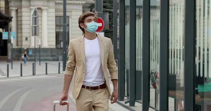 Young businessman in medical mask and formal clothing walking outside of railway station with luggage. Concept of business trip, protection and health care.