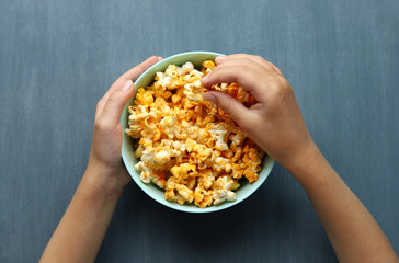 Blue dish full of popcorn on a gray background. A child tries popcorn. Selective focus. Hands and popcorn