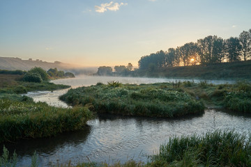 Fototapeta na wymiar Summer time. Morning dawn over the river in a hazy, thoughtful haze. Beautiful view of the forest and river covered with fog early in the morning.