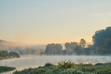 Summer time. Morning dawn over the river in a hazy, thoughtful haze. Beautiful view of the forest and river covered with fog early in the morning.