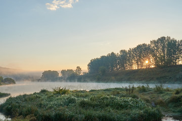 Summer time. Morning dawn over the river in a hazy, thoughtful haze. Beautiful view of the forest and river covered with fog early in the morning.