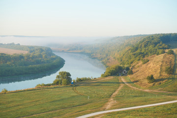 Fototapeta premium Summer time. Morning dawn over the river in a hazy, thoughtful haze. Beautiful view of the forest and river covered with fog early in the morning.