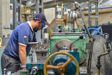 Working turner wearing a protective antiviral mask on a lathe in a workshop. The machine operator at the workplace controls the equipment.