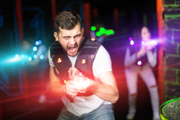 Emotional guy playing laser tag with friends on dark labyrinth in colorful beams of laser pistols