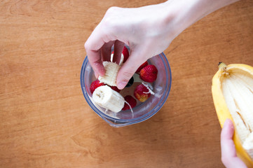 Hands put banana slices in a blender with strawberries, top view