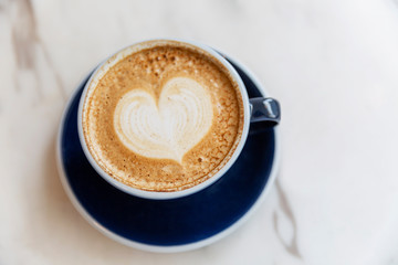 Cappuccino with heart shaped foam in a blue cup on a marble table surface. Top view.
