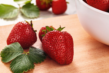 Ripe strawberries and leaves on wooden board near bowl.