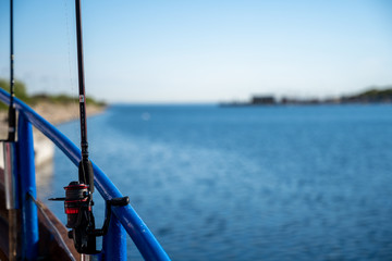 A backlit closeup picture of a blue rail and a fishing rod on a boat. Clear blue sky and ocean in the background