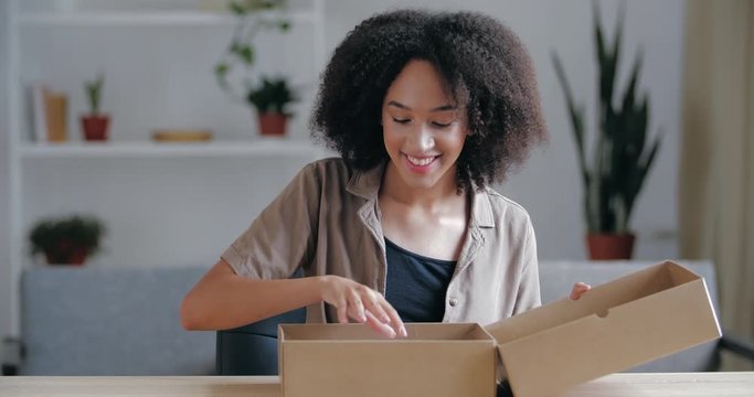 Close up curly haired young brunette receives parcel from courier from internet store, accepts online ordered product, rejoices, claps her hands. Happy customer opens unpacks carton with goods at home