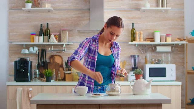 Young woman preparing green tea for breakfast in a modern kitchen using teapot sitting near the table. Putting with hands, pressing herbal, healthy, tea leaves, in pot, in the morning.