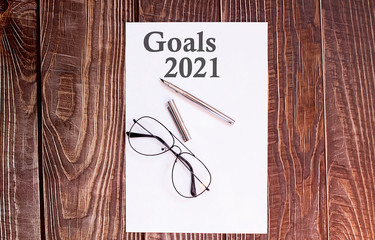 GOALS 2021 words written on a white piece of paper laid on a wooden table with pen and glasses. Top view, concept in business.